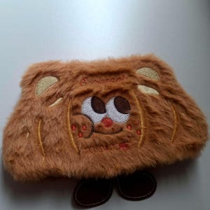 K&ouml;nnte beinhalten: Eine braune, pl&uuml;schige, rechteckige Tasche mit einem aufgestickten Cartoon-B&auml;rengesicht auf der Vorderseite. Der B&auml;r hat wei&szlig;e Augen, eine rote Nase und einen l&auml;chelnden Mund. Die Tasche hat zwei braune, oval geformte F&uuml;&szlig;e.