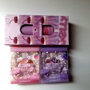 Puede incluir: Un conjunto de productos de maquillaje, que incluye un stick de rubor y dos compactos de resaltador de diamantes. El stick de rubor está en una caja rosa con un diseño de cereza. Los compactos están etiquetados como "Juicy Peach" y "Grape Jelly".