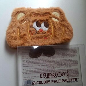 K&ouml;nnte beinhalten: Eine braune, flauschige Make-up-Tasche mit einem gestickten Cartoon-B&auml;rengesicht. Die Tasche hat eine rechteckige Form mit abgerundeten Ecken und einem Rei&szlig;verschluss. Der Text "KEVIN & COCO" und "12 COLORS FACE PALETTE" sind auf der Verpackung zu sehen.