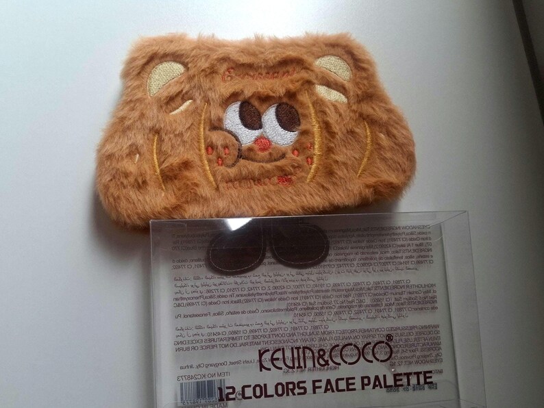 K&ouml;nnte beinhalten: Eine braune, flauschige Make-up-Tasche mit einem gestickten Cartoon-B&auml;rengesicht. Die Tasche hat eine rechteckige Form mit abgerundeten Ecken und einem Rei&szlig;verschluss. Der Text "KEVIN & COCO" und "12 COLORS FACE PALETTE" sind auf der Verpackung zu sehen.