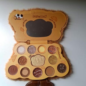 K&ouml;nnte beinhalten: Hellbraune Lidschattenpalette in B&auml;renform mit 12 Farben und einem Spiegel. Die Palette tr&auml;gt die Aufschrift "KEVIN&COCO" und "Face Palette". Das Wort "Croissant" ist ebenfalls sichtbar.