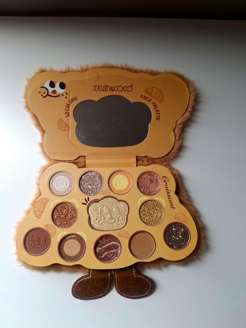 K&ouml;nnte beinhalten: Hellbraune Lidschattenpalette in B&auml;renform mit 12 Farben und einem Spiegel. Die Palette tr&auml;gt die Aufschrift "KEVIN&COCO" und "Face Palette". Das Wort "Croissant" ist ebenfalls sichtbar.
