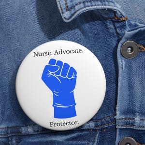 May include: A white button with the words "Nurse. Advocate. Protector." in black text. A blue fist graphic is in the center. The button is pinned to a blue denim jacket.
