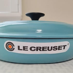 May include: A teal Le Creuset Dutch oven with a black handle on the lid. The oval-shaped pot has two side handles and a white sticker with the brand name. The pot is on a white surface.