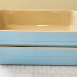 Puede incluir: Dos fuentes de horno rectangulares apiladas una sobre otra. Cada fuente presenta un exterior azul claro con un acabado liso y brillante y un interior de color crema. Los lados tienen estrías horizontales y cada extremo incluye una pequeña asa.
