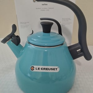 May include: A teal Le Creuset kettle with a black handle and spout. The kettle has a rounded body and a black lid. The brand name is displayed on the front. The box behind the kettle says "kettles".