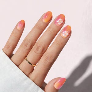 May include: A hand displaying oval-shaped nails with an orange and pink gradient. Two nails are decorated with white flower designs. A gold ring adorns the ring finger. The nails offer a vibrant and stylish aesthetic.