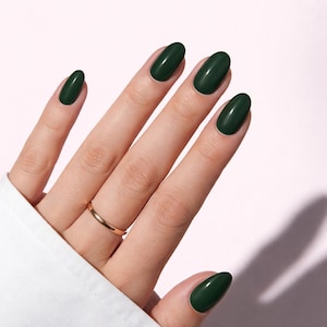 Puede incluir: Una mano con uñas ovaladas pintadas de un verde bosque oscuro. Un anillo de oro adorna el dedo anular. Las uñas son brillantes y reflejan la luz. El fondo es de color rosa pálido.