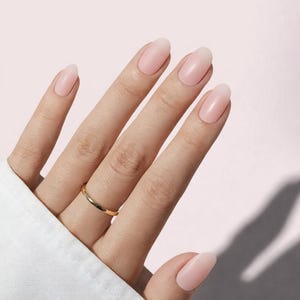 May include: A hand with oval-shaped, pale pink nails and a gold ring on the ring finger. The nails have a natural, glossy finish. The background is a soft pink.