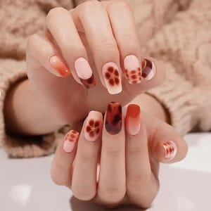 May include: A close-up of a hand with a manicure featuring various nail designs. The nails showcase a range of colors, including pink, orange, and brown, with patterns like tortoiseshell and floral accents. The nails are of varying lengths and shapes.