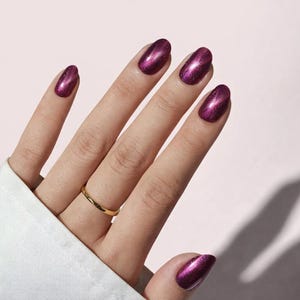 May include: A hand with polished nails in a deep magenta color, with a metallic sheen. The nails are oval-shaped. A gold ring is on the ring finger. The background is a soft pink.