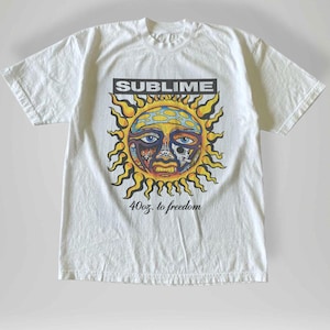 Puede incluir: Camiseta blanca con un gráfico de sol colorido, la palabra "SUBLIME" en un recuadro negro y el texto "40oz. to freedom". El diseño del sol incluye detalles amarillos, azules, naranjas y negros. Una camiseta gráfica informal.