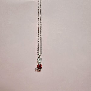 Puede incluir: Collar con colgante de gato plateado con cuerpo de gema roja y cabeza de gema transparente. El collar cuelga de una delicada cadena sobre un fondo rosa. El gato está sentado.