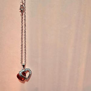 Könnte beinhalten: Silberfarbene Halskette mit einem Anhänger aus zwei Herzen. Eines ist ein rotes Herz, das andere ein offenes Herz mit kleinen, klaren Kristallen. Die Kette ist zart und vor einem rosa Hintergrund angeordnet.