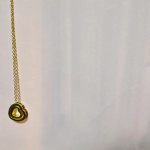 Puede incluir: Un collar dorado con un colgante en forma de corazón. El colgante tiene un corazón más pequeño en su interior y está adornado con pequeñas piedras preciosas rojas. La cadena es delicada y también dorada.