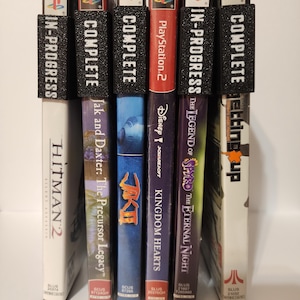 Puede incluir: Una colección de carátulas de videojuegos de PlayStation 2, incluyendo "Hitman 2" y "Kingdom Hearts". Cada carátula está etiquetada con una etiqueta negra que indica el estado del juego, "Complete" o "In-Progress".