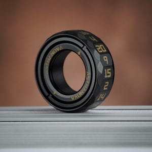 Può includere: Un anello nero e oro con la scritta "ASTRANOVA" e numeri. L'anello ha un bordo esterno sfaccettato con numeri dorati. L'anello è su una superficie argentata con sfondo marrone.