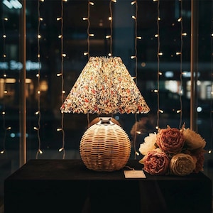 Può includere: Una lampada da tavolo decorativa con base in vimini intrecciato e paralume a fantasia floreale. La lampada è posizionata su una superficie nera, con un bouquet di fiori a destra. Luci a catena visibili sullo sfondo.
