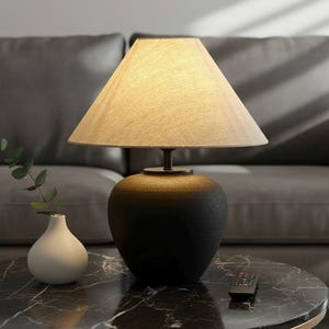 Puede incluir: Una lámpara de mesa negra con una pantalla beige sobre una mesa con tapa de mármol. La lámpara tiene una base redondeada y una pantalla cónica. Un pequeño jarrón blanco con vegetación y un mando a distancia también están sobre la mesa.