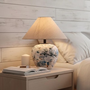 Peut inclure: Une lampe de chevet avec un abat-jour beige et un pied décoratif avec des accents bleus, blancs et bruns. Une bougie blanche, un livre ouvert et une montre-bracelet sont sur la table de chevet en bois clair.
