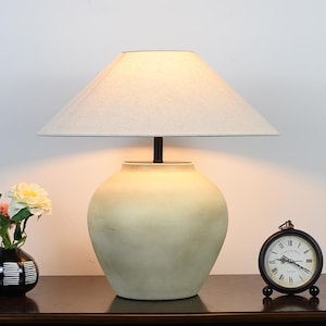 May include: A cream-colored table lamp with a beige lampshade. The lamp has a rounded base and a dark brown stem. A small black clock and a vase of flowers are on the table.