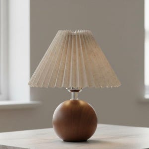 May include: A small table lamp with a pleated beige lampshade and a round, dark brown wooden base. The lamp has a silver-colored stem and sits on a light-colored surface. The lamp is designed for indoor use.
