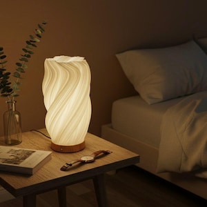 May include: A cream-coloured, swirled ceramic table lamp with a wooden base sits on a wooden bedside table. A watch and a book are also on the table. A glass vase with greenery is to the left of the lamp. The lamp is lit, casting a warm glow.