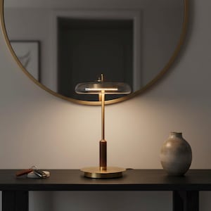 May include: A contemporary brass table lamp with a clear glass shade and a dark wood detail. The lamp sits on a dark table with a vase and keys. A gold-framed mirror is in the background.