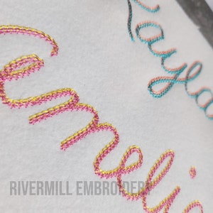 Pu&ograve; includere: Primo piano di due nomi ricamati, "Amelia" e "Kayla", cuciti con filo rosa e oro su uno sfondo di tessuto bianco.
