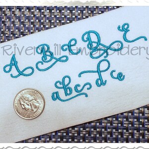 May include: A close-up of a white fabric with turquoise blue embroidered letters. The letters are in a cursive font and include the letters A, B, C, D, and E. The letters are arranged in a random order. The fabric is on a woven background.