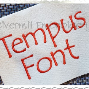 May include: White fabric with the words "Tempus Font" embroidered in red. The text is in a cursive style. The words "Rivermill Embroidery" are in the upper left corner.