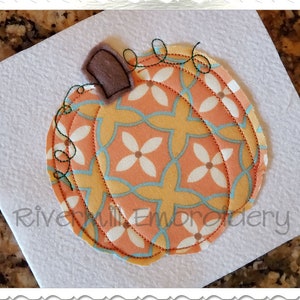 May include: A close-up of a felt pumpkin applique with a brown stem and a peach, yellow, and teal patterned fabric. The pumpkin is stitched with a decorative stitch.