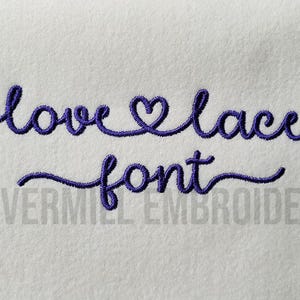May include: Embroidered font design that says "love lace font" with a heart in place of the "o" in love. The text is in a cursive style and is embroidered in purple thread on a white background.