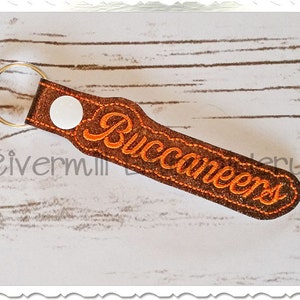 Puede incluir: Un llavero de cuero marrón con un botón blanco y la palabra "Buccaneers" bordada en naranja.