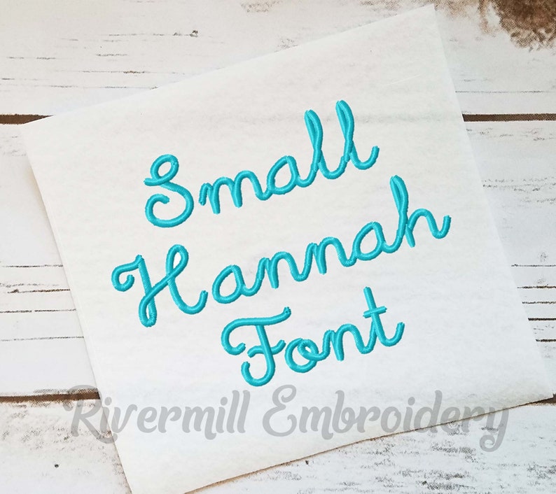 K&ouml;nnte beinhalten: Nahaufnahme eines wei&szlig;en Stoffes mit dem Text "Small Hannah Font", der in t&uuml;rkisblauer kursivschrift gestickt ist. Der Text "Rivermill Embroidery" ist ebenfalls in der linken unteren Ecke sichtbar.