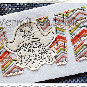May include: A white felt applique of the word "LOVE" with a pirate character in the center. The letters are made of fabric with a chevron pattern in orange, brown, and blue.