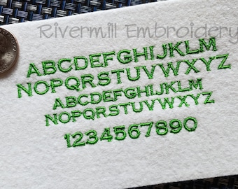 May include: A white fabric swatch with green embroidered letters and numbers. The letters are in two rows, with the first row being A-Z and the second row being N-Z. The numbers are 1-9 and 0.
