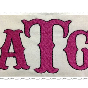 Może przedstawiać: Naszywka haftowana z literami ATG w różowym kolorze z czarnymi konturami na białym tle z tkaniny.