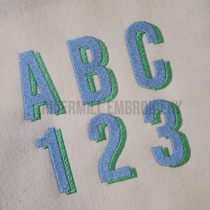 以下が含まれることがあります： 青と緑の配色で刺繍されたアルファベットのA、B、Cと数字の1、2、3。文字は緑色のアウトラインで、青色で塗りつぶされています。