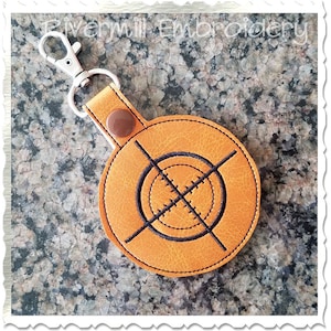 May include: A brown leather keyring with a black embroidered target design. The keyring has a silver metal ring and a brown button.