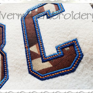 Puede incluir: Dos letras bordadas, "3" y "C", en un patr&oacute;n de camuflaje marr&oacute;n y blanco con costuras azules. Las letras est&aacute;n sobre un fondo blanco.
