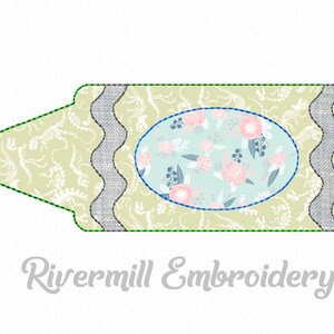 May include: A green crayon with a dinosaur pattern and a light blue oval with a floral pattern in the center. The crayon has a wavy edge and a gray border. The text "Rivermill Embroidery" is below the crayon.