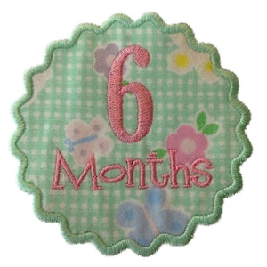 May include: A green and white fabric circle with a pink embroidered number 6 and the text "Months". The circle has a scalloped edge and a floral pattern.