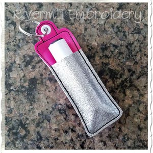 May include: A silver glitter keychain holder with a pink flap and a black zipper. The holder is designed to hold a lip balm or other small item.