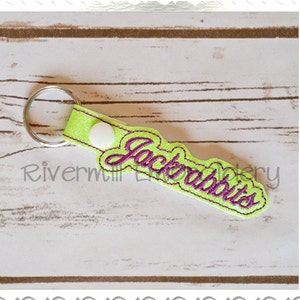 May include: A neon green and purple keychain with the word "Jackrabbits" embroidered in purple script. The keychain has a silver key ring and a white snap closure. The background is a light wood grain.