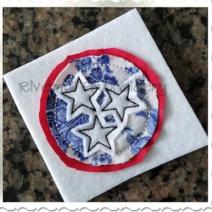 May include: A white felt circle with a red border and a blue and white fabric circle with three white stars inside. The fabric circle has a blue floral pattern.