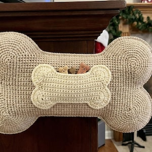 May include: A beige, crocheted dog bone-shaped stocking with a smaller bone-shaped pocket. The pocket contains several dog treats. A loop is attached for hanging. The stocking is displayed near a fireplace.