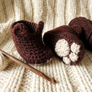 May include: Handmade crochet baby booties in dark brown with cream-colored paw prints. A wooden crochet hook and a ball of cream-colored yarn are next to the booties. A skein of dark brown yarn sits in the background, all on a cream-colored knitted blanket.