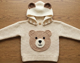 May include: Cream-colored, crocheted baby hoodie with a bear face applique. The hoodie features a hood with bear ears, wooden buttons, and long sleeves. The bear face is brown with a black nose and is centered on the front.