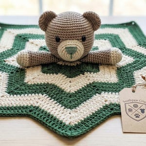 May include: A handmade crochet teddy bear with a green nose and a matching green and cream-coloured star-shaped blanket. A ball of yarn, a crochet hook, and a tag with a logo are also present. The bear is lying on the blanket.